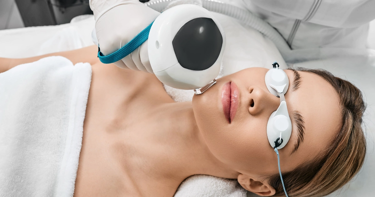 High-angle close-up of a woman receiving IPL spot treatment, wearing protective goggles, draped in white towels, in a bright clinical setting in Pueblo, CO